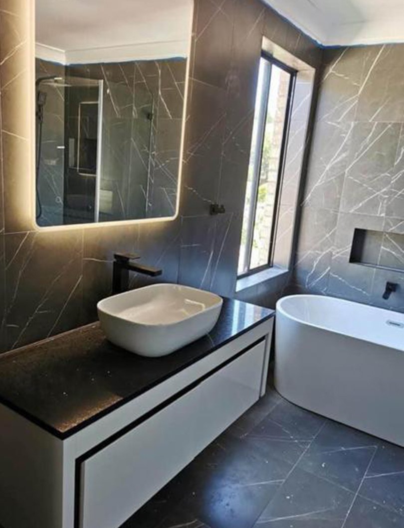Bathroom renovation with dark marble-look tiling, a backlit mirror, a white vessel basin on a black stone benchtop and a freestanding bath beside a garden window